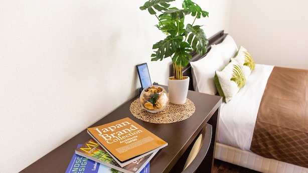 A neatly arranged hotel room with a bed, a bedside table with books, a potted plant, and a small decorative item. - Mr. Kinjo Violette Kukomae (Naha)