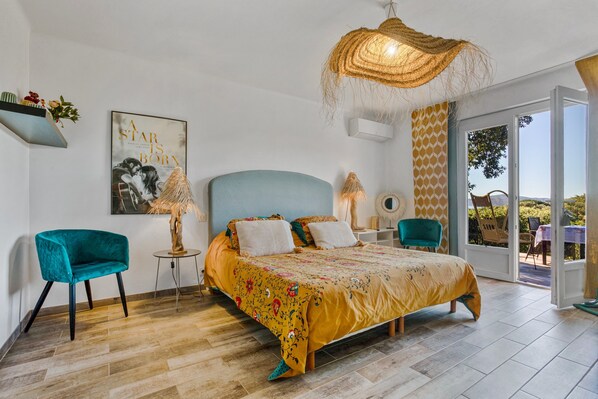 A bedroom with a bed, a blue chair, a small table, a hanging light fixture, and a view through an open door.
