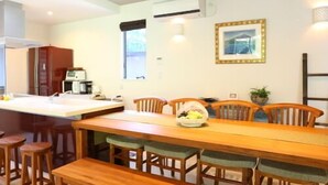 Een eetgedeelte met een houten tafel en stoelen, een bank, een keukenhoek met een koelkast, en een raam met zonwering.