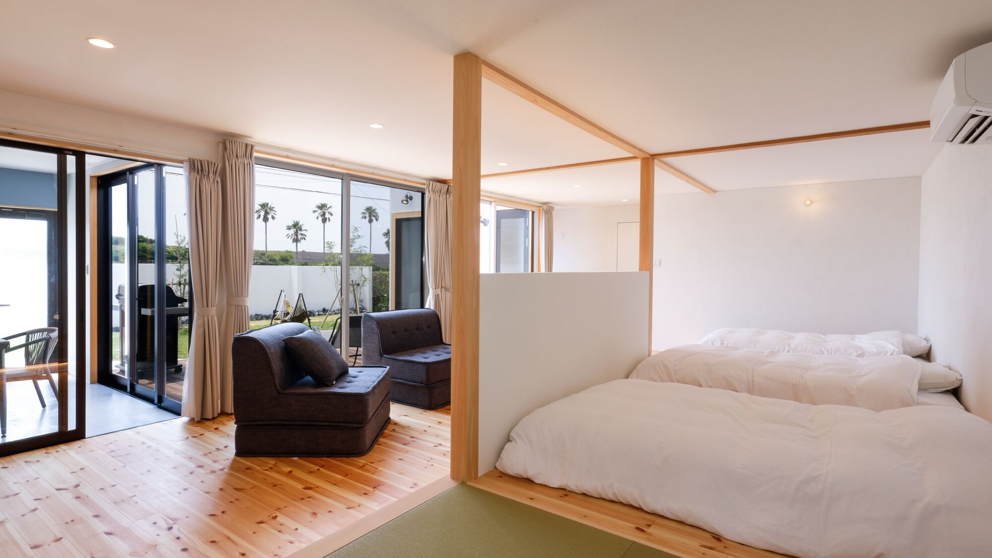 A bedroom with a large bed, a seating area with two chairs, and a sliding glass door leading to an outdoor area with a view of palm trees.