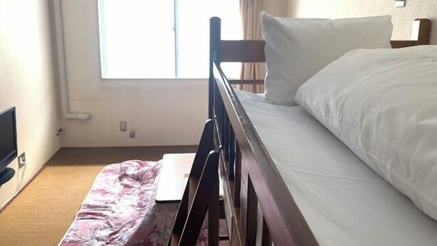 Una cama individual con estructura de madera y ropa de cama blanca en una habitación con ventana, escritorio y televisor.
