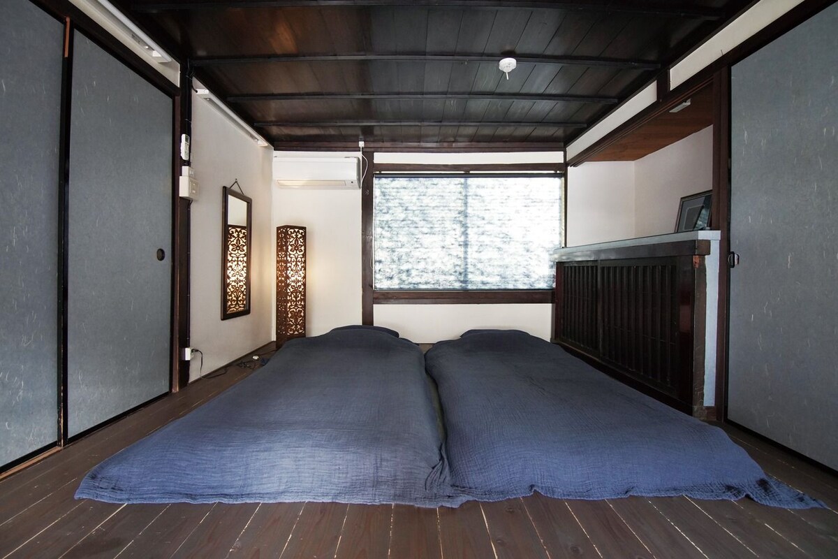 A room with a wooden ceiling, a window with a view of trees, a radiator, and a bed with a blue blanket.