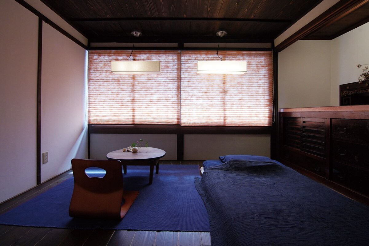 A small, cozy room with a bed, a table, and a chair. There is a window with blinds, a ceiling light, and a dresser.