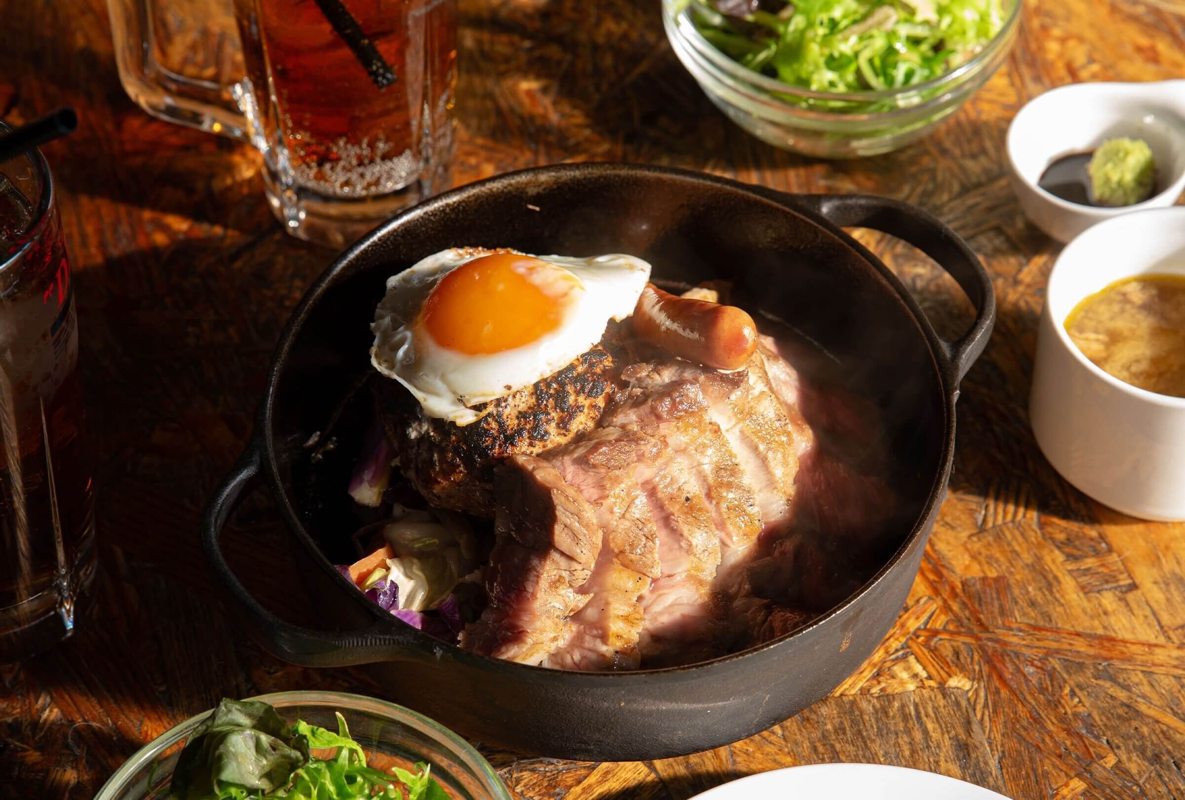 Comida con jamón a la plancha y huevo en una sartén de hierro fundido, acompañada de ensalada y bebidas sobre una mesa de madera.