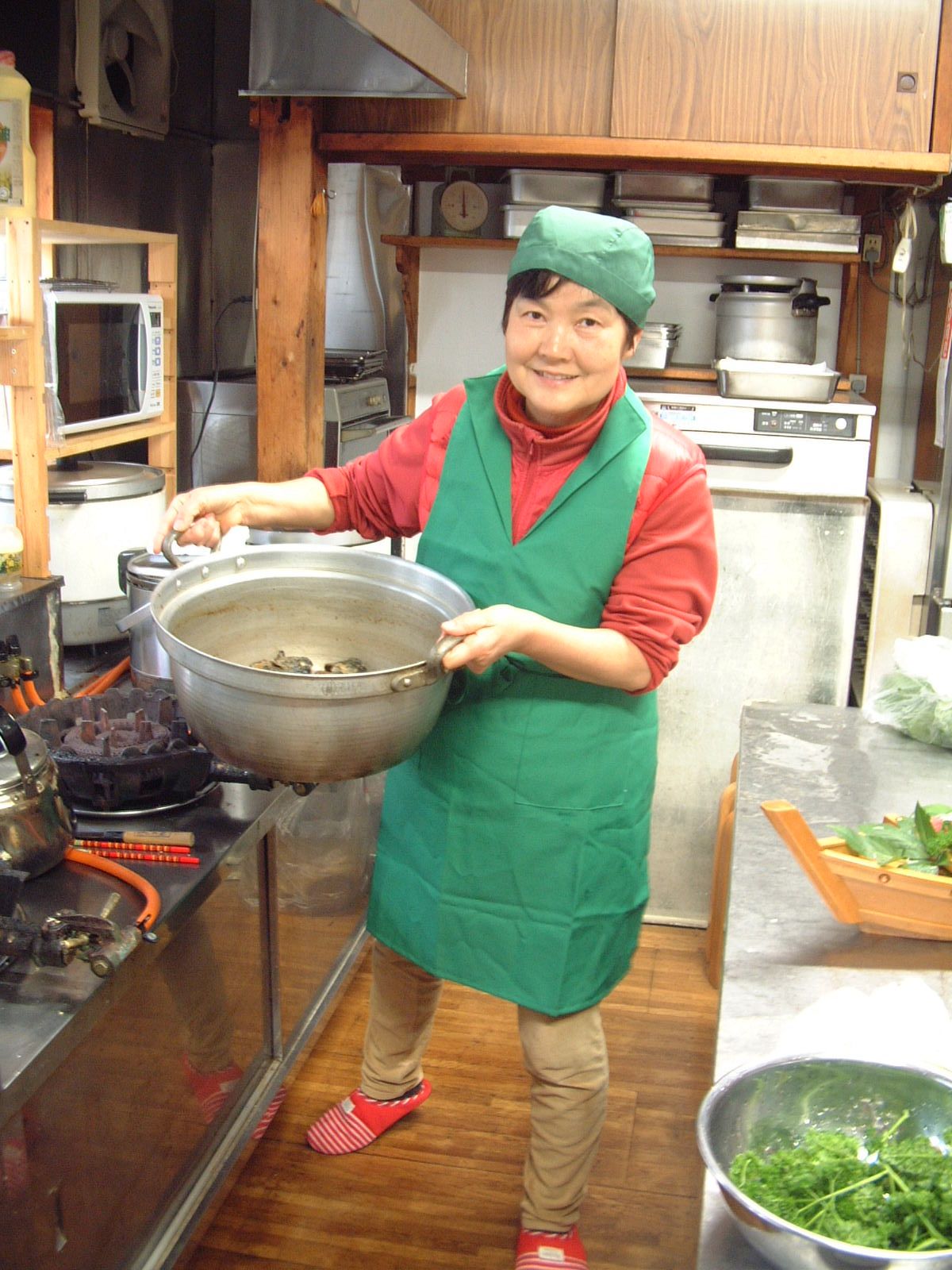 Una persona en una cocina sosteniendo una olla grande, con varios electrodomésticos y utensilios de cocina a la vista.