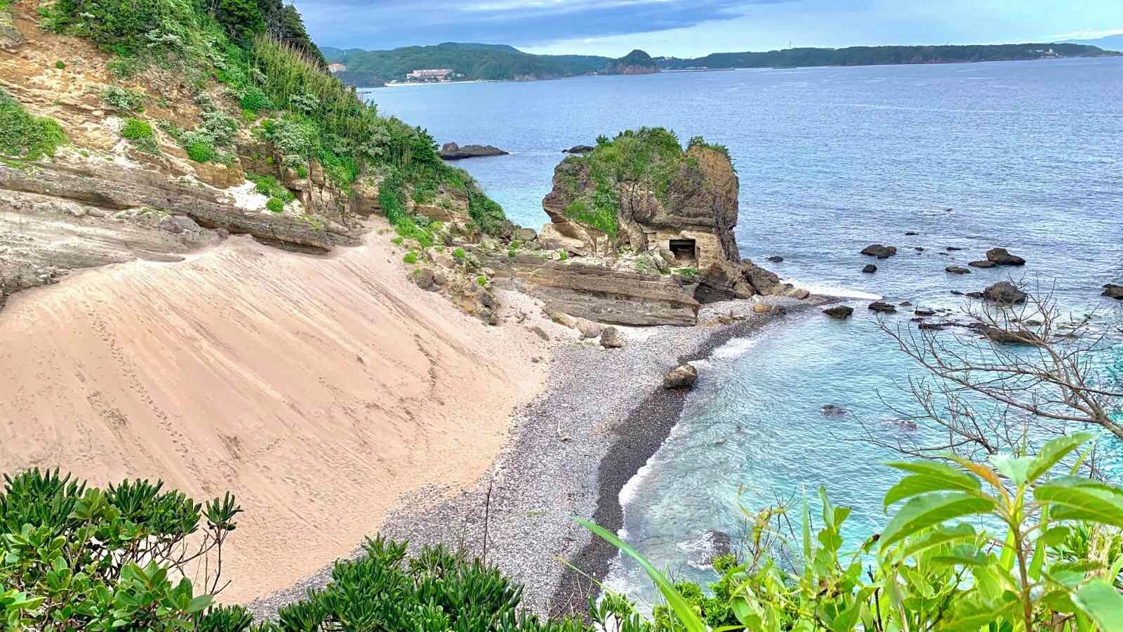 Paisagem costeira com praia de areia, águas azuis cristalinas e um afloramento rochoso coberto de vegetação.