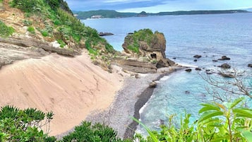Paisagem costeira com praia de areia, águas azuis cristalinas e um afloramento rochoso coberto de vegetação.