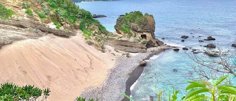 A coastal landscape with a sandy beach, clear blue waters, and a rocky outcrop with vegetation.