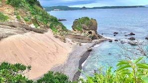 Paisagem costeira com praia de areia, águas azuis cristalinas e um afloramento rochoso coberto de vegetação.