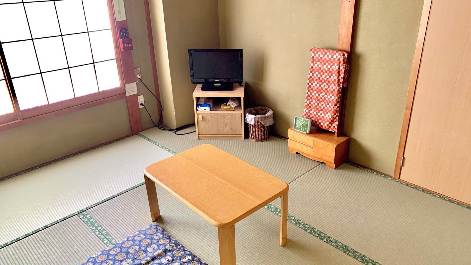 Una habitación con suelo de tatami, una mesa de madera, un televisor sobre un mueble bajo, un armario pequeño y un biombio japonés tradicional.