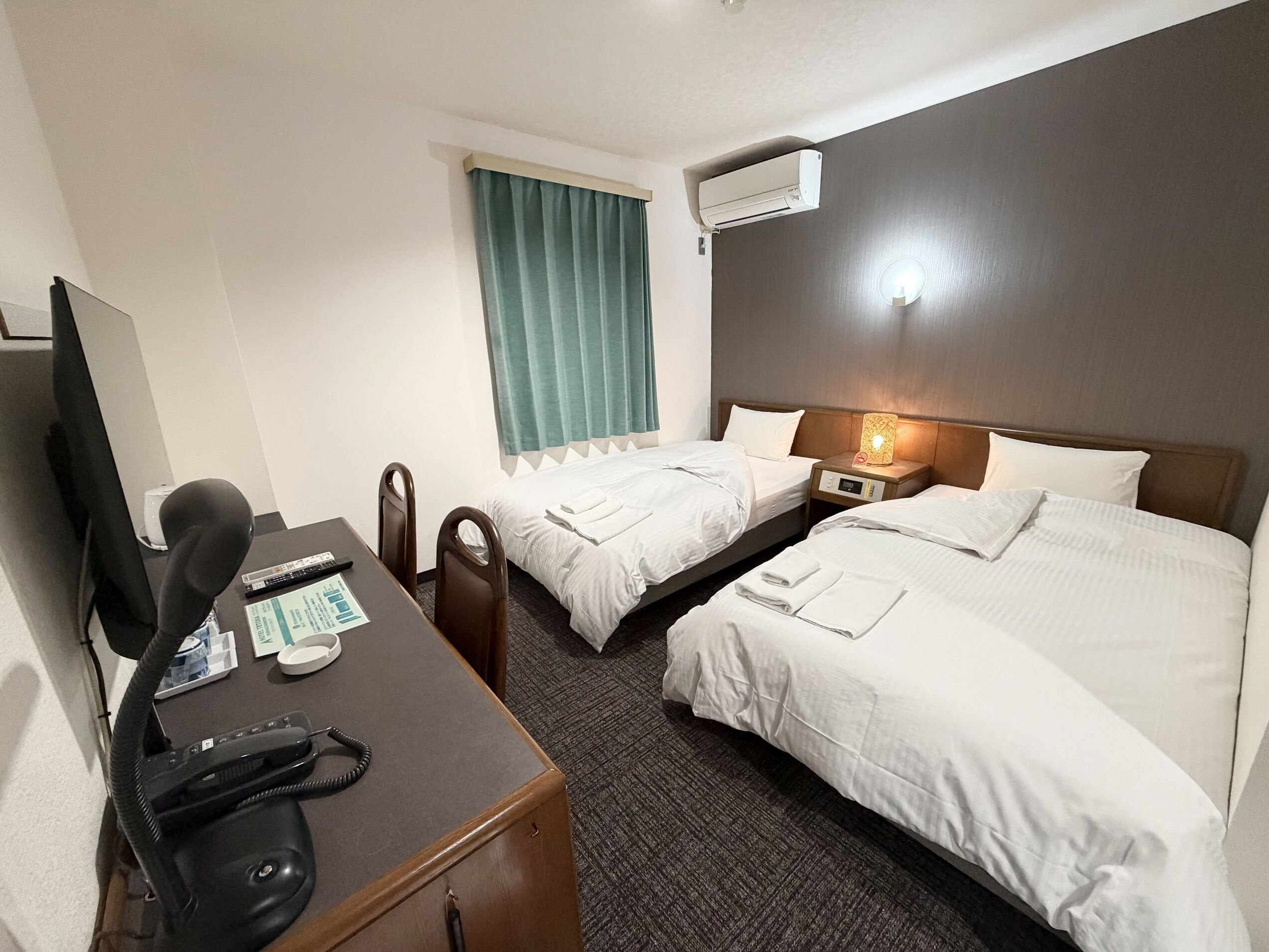 A hotel room with two beds, a desk with a telephone, a television, and an air conditioning unit.