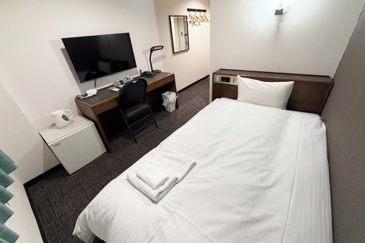 A hotel room with a bed, a desk, a chair, a television, a mirror, and a wall-mounted light fixture.