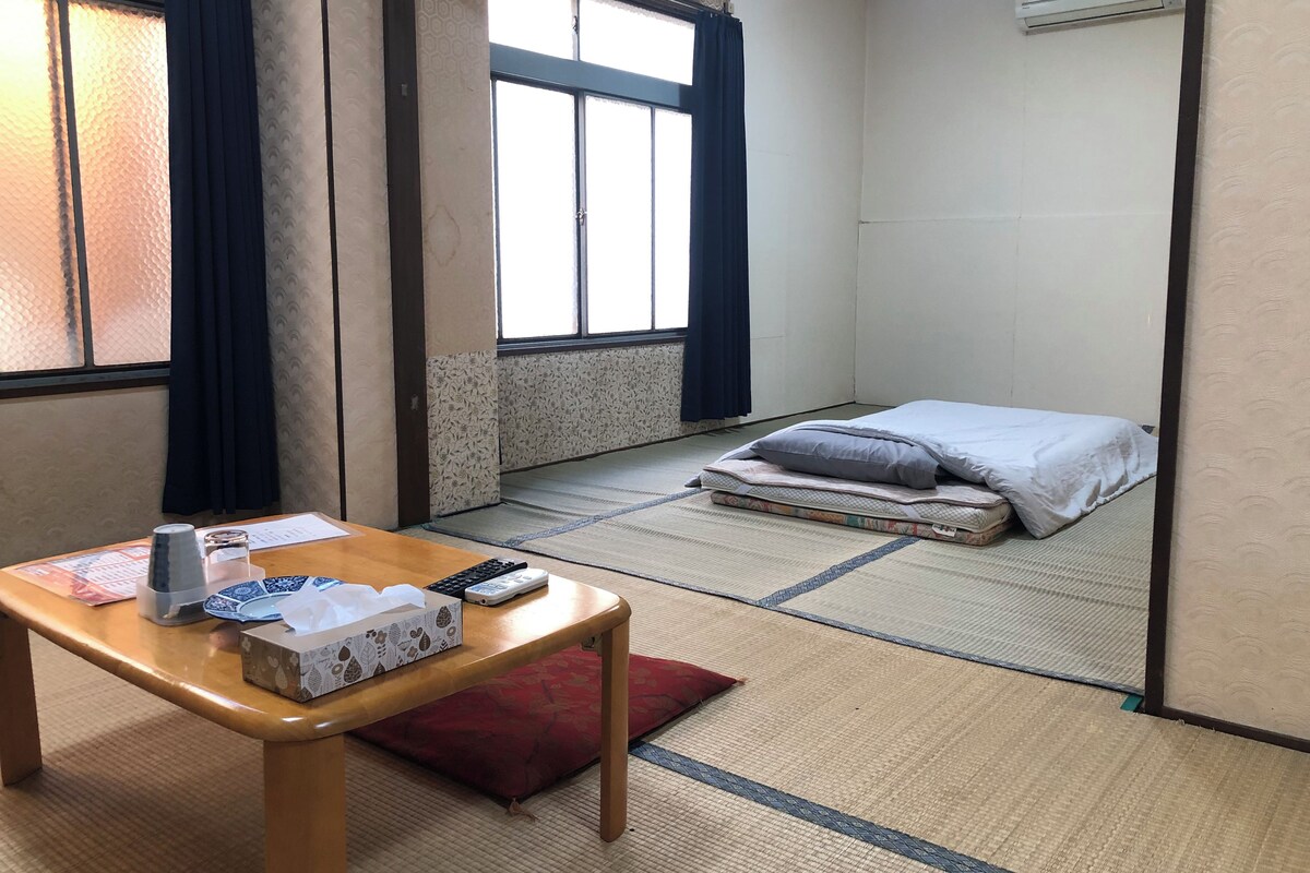 A room with tatami flooring, a bed with a white and grey duvet, a wooden table with a tissue box, a remote control, and a small red pillow.