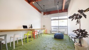Ein Zimmer mit grünem Kunstrasenteppich, einem Holztisch mit einem Laptop und bunten Stühlen.