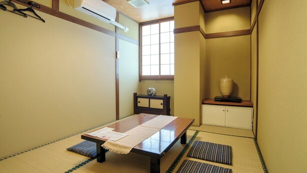 A traditional Japanese room with tatami flooring, a low table with a placemat, a window with a view, and a cabinet with a vase. - Traveler's Inn Takagi (Matsumoto)