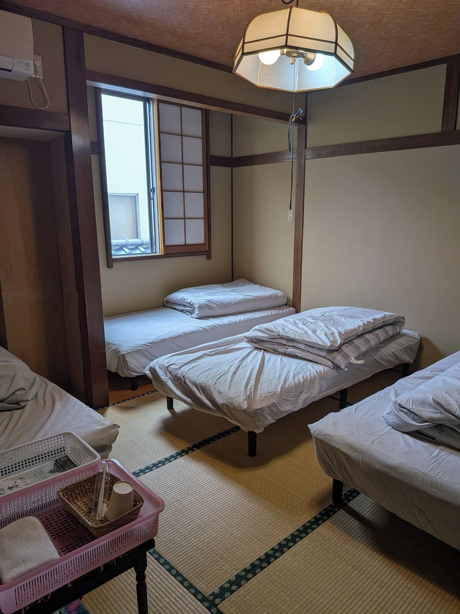 A room with two single beds, a small table with a basket, a window with a view, and a ceiling light fixture.