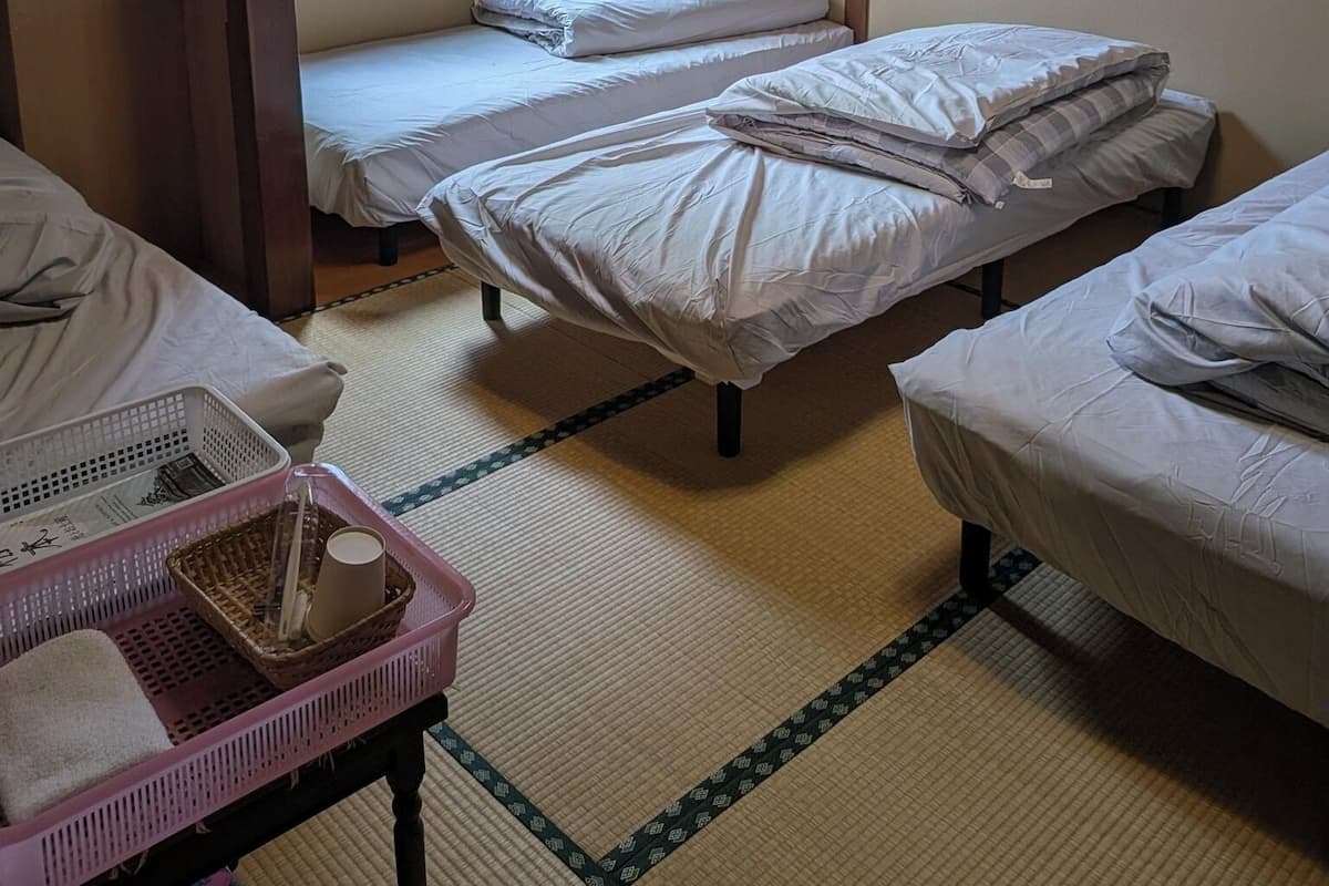 A room with two single beds, a small table with a basket, a window with a view, and a ceiling light fixture.