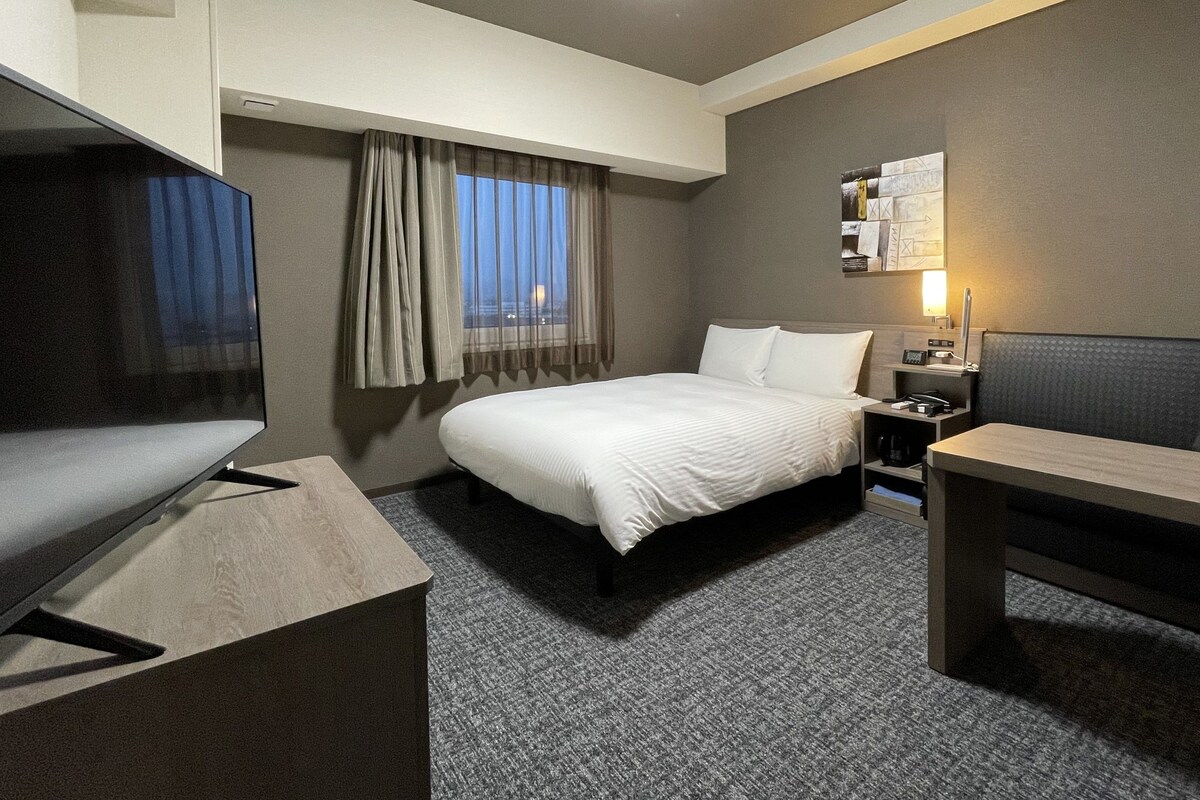 A hotel room with a bed, a desk, a chair, a TV, and a window with curtains.