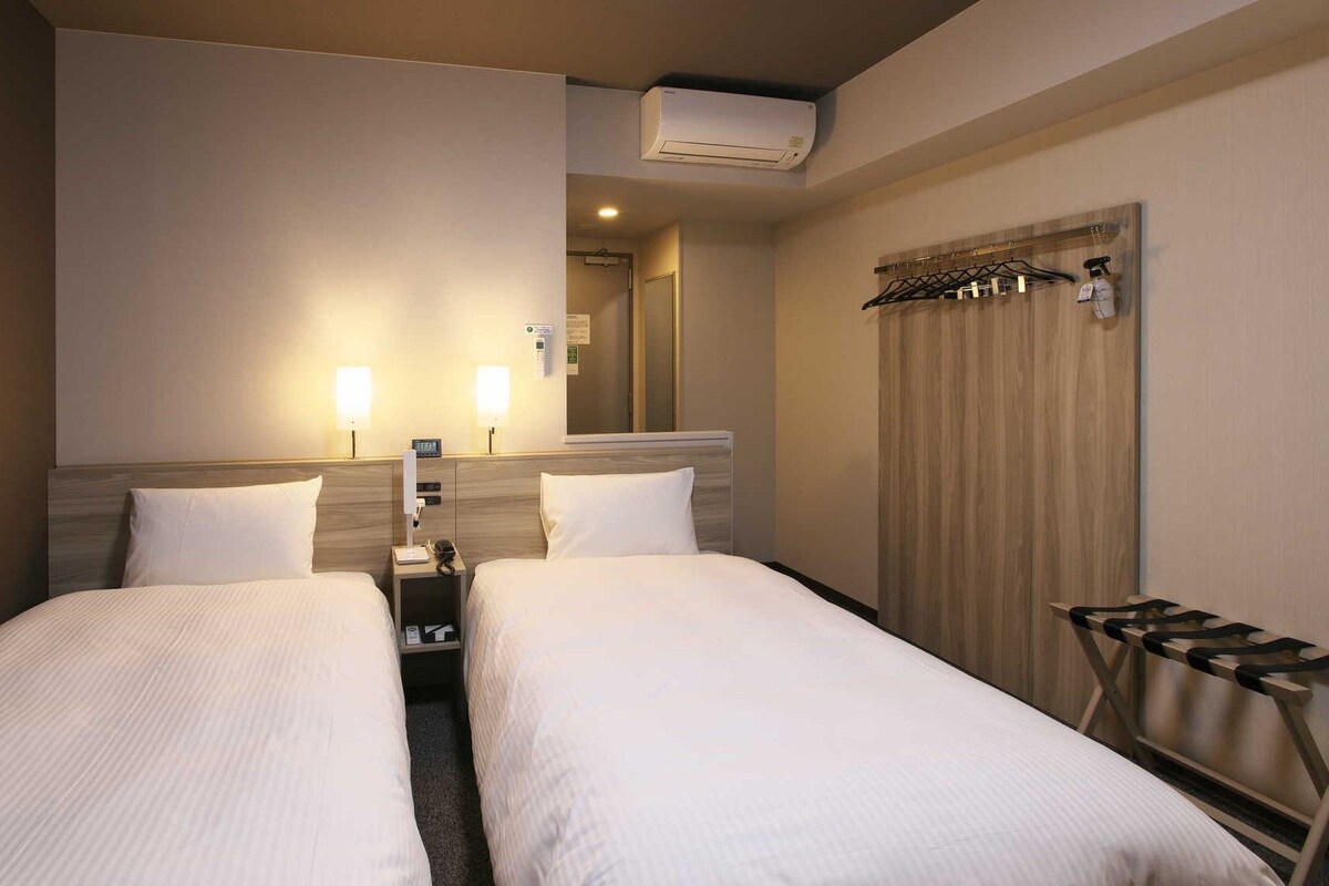 A hotel room with two single beds, a wooden headboard, a mirror, a coat rack, and an air conditioning unit.