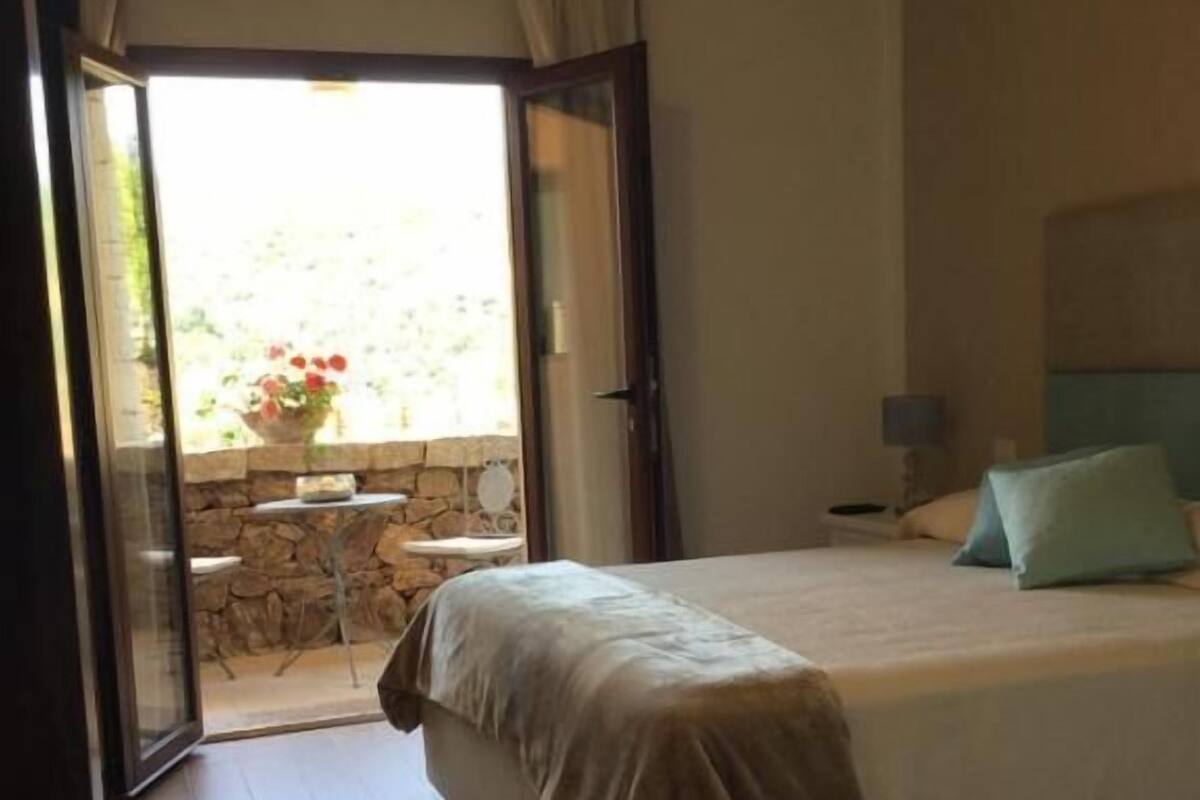 A bedroom with a bed, a nightstand, a lamp, and a view of a garden through an open door.