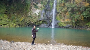 A person standing by a clear, rocky lake with a waterfall in the background. - Guest House Amigo (Toyooka)