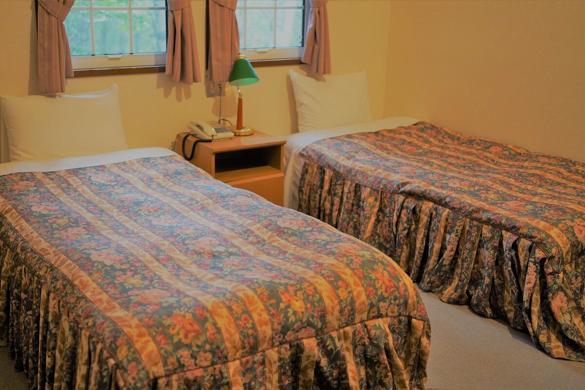A room with two single beds, a nightstand with a lamp, and a window with curtains.