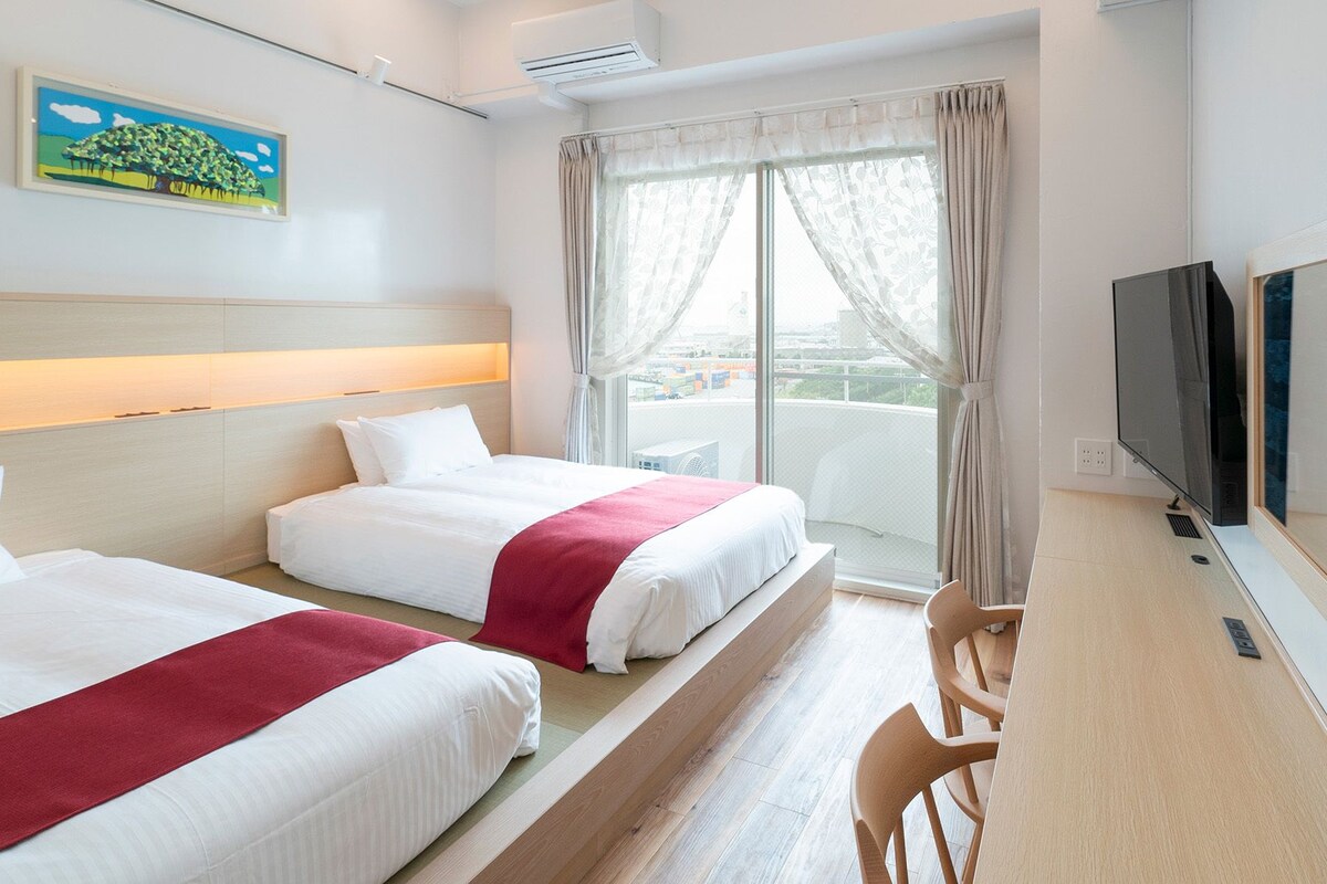 Habitación de hotel con dos camas, televisión, escritorio y un ventanal grande con cortinas.