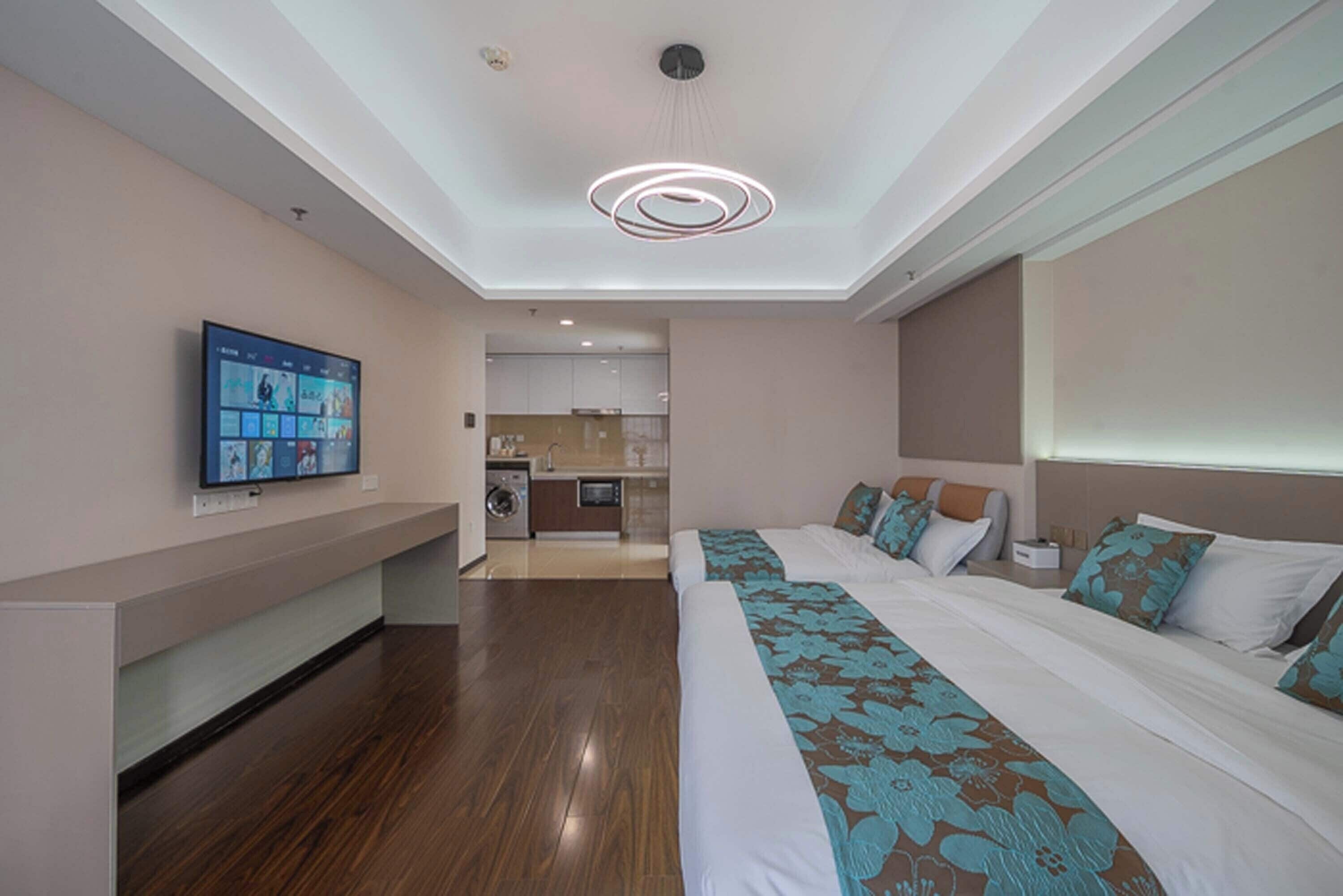 Habitación de hotel con dos camas, un televisor fijado a la pared, una lámpara de techo moderna y vista al área de la cocina.