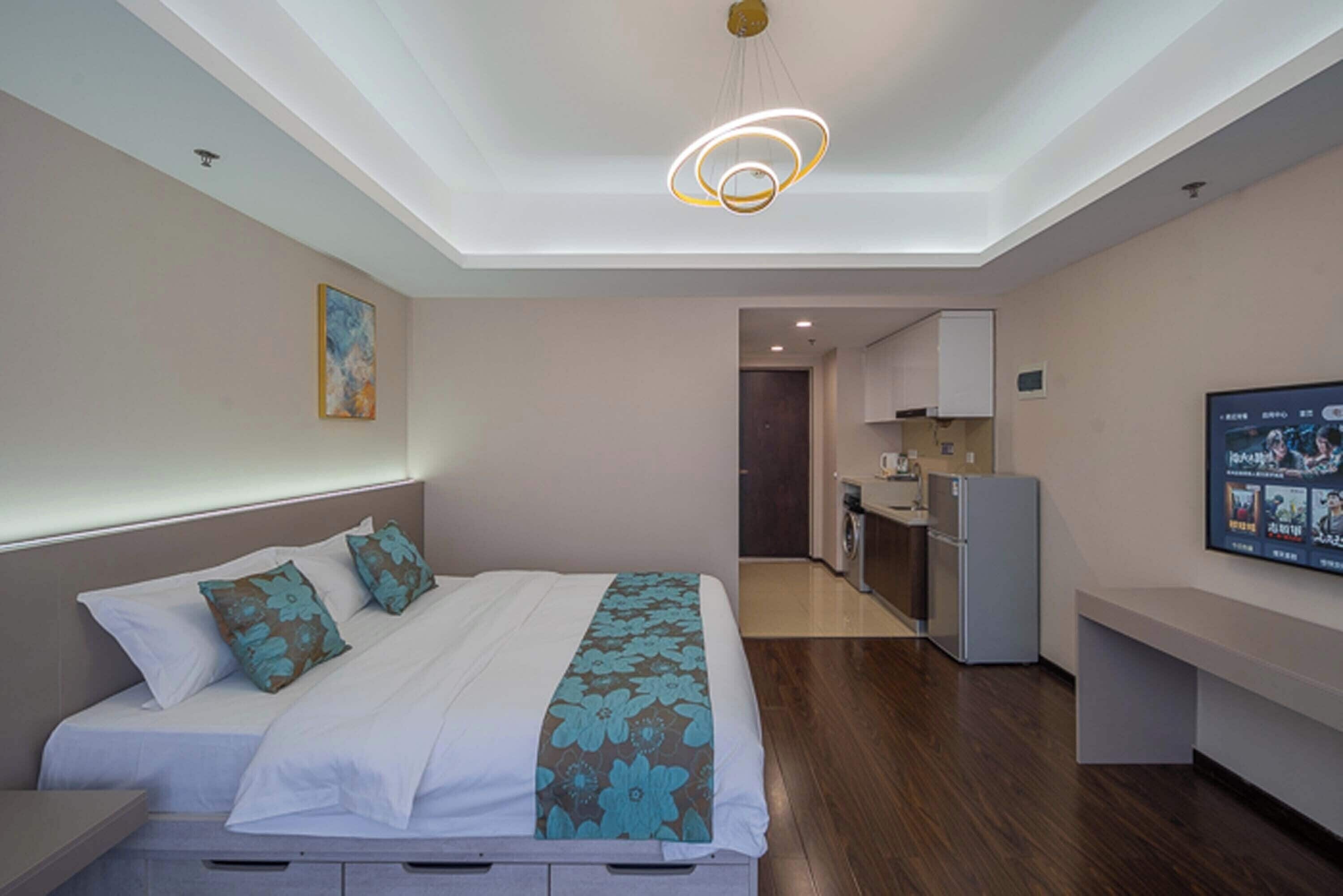A modern hotel room with a neatly made bed, a small kitchenette, and a television on the wall.