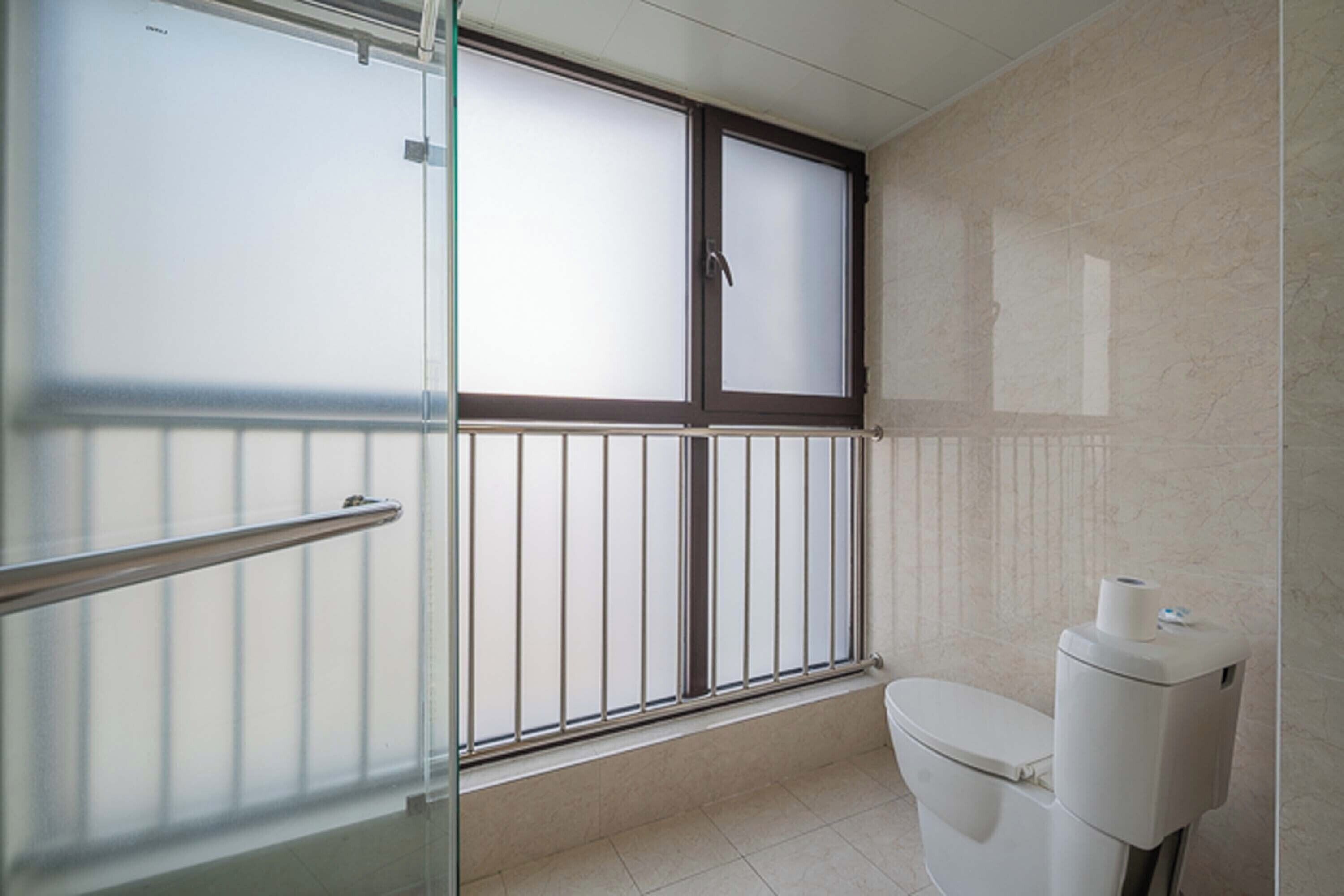 A bathroom with a toilet, a window with blinds, and a glass door with a metal handle.