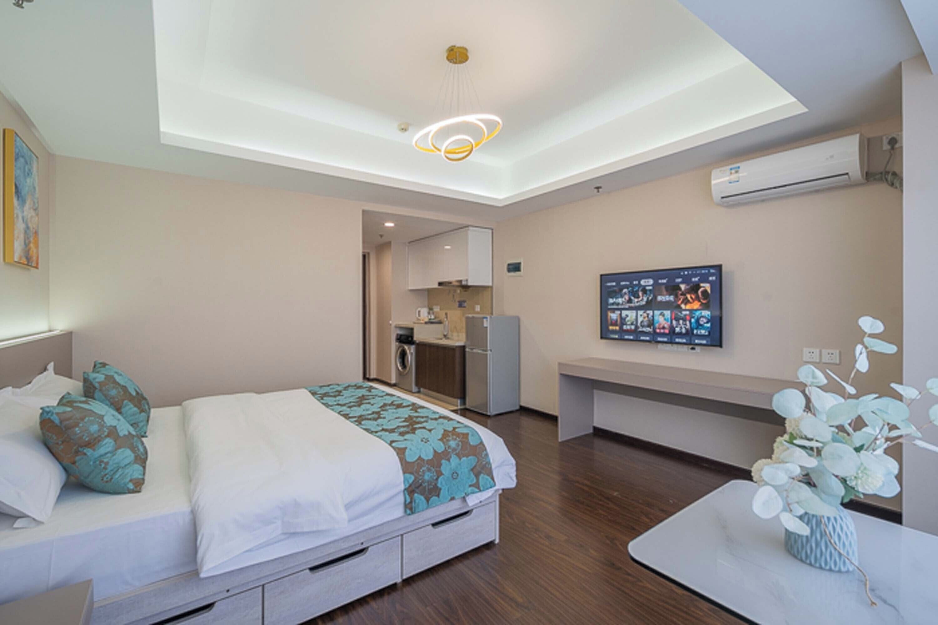 A modern hotel room with a bed, a desk, a small kitchenette, and a ceiling light fixture.