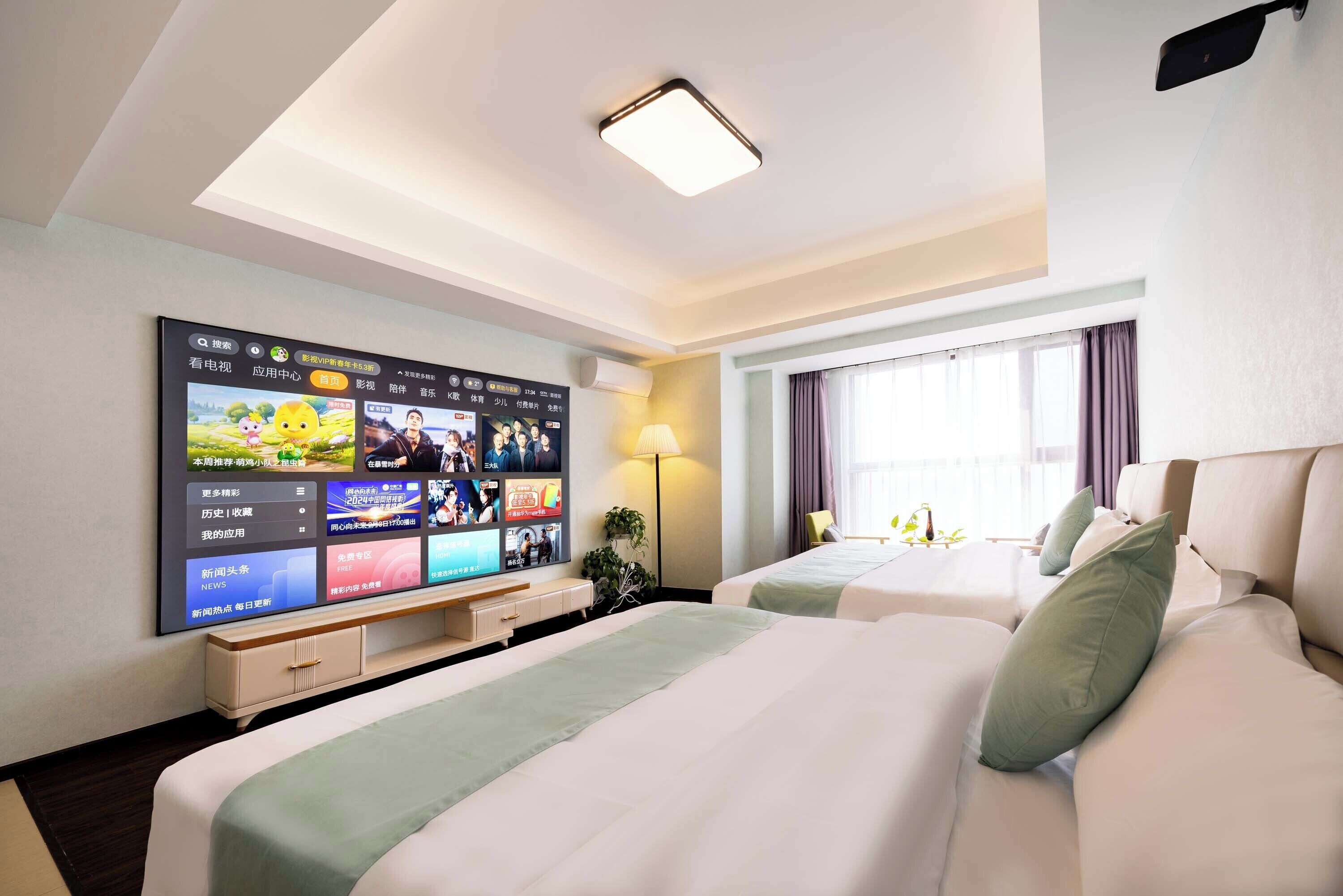 A hotel room with a large flat-screen TV mounted on the wall, two beds with green pillows, a bedside table, a lamp, and a window with curtains.