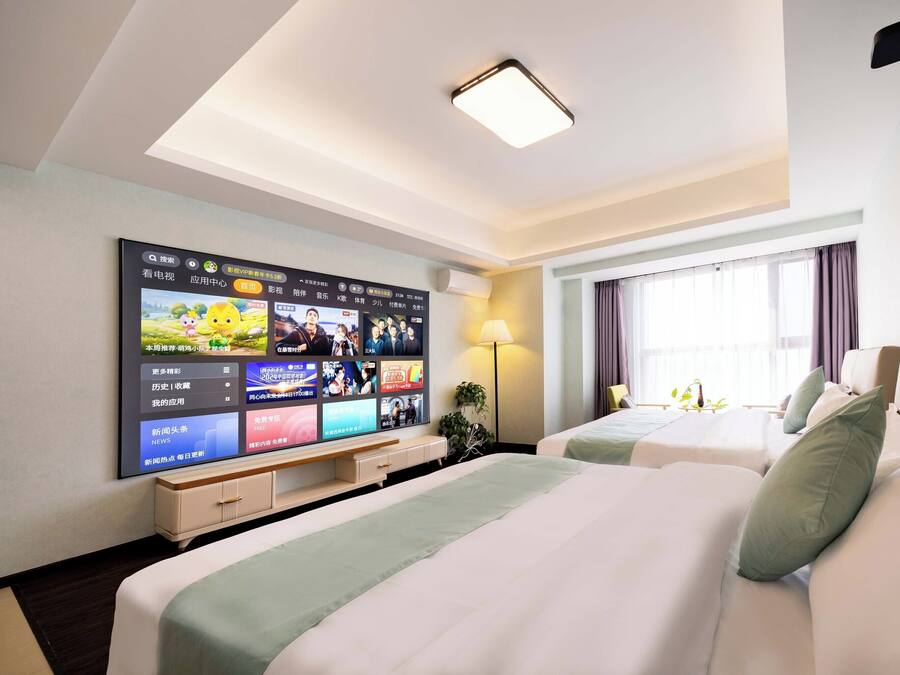 Habitación de hotel con un televisor de pantalla plana grande montado en la pared, dos camas con almohadas verdes, una mesita de noche, una lámpara y una ventana con cortinas.