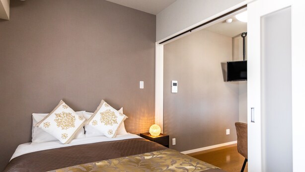 A bedroom with a bed, pillows, bedside table, lamp, and a TV mounted on the wall. - Mr.kinjo in Arcade Ange Miona (Naha)