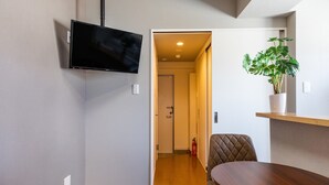 A modern room with a flat-screen TV mounted on the wall, a wooden dining table with a single chair, and a potted plant on a shelf. - Mr.kinjo in Arcade Ange Miona (Naha)