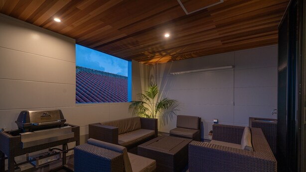 Un área de estar al aire libre con muebles de mimbre, una parrilla y una gran pantalla que muestra un atardecer.
