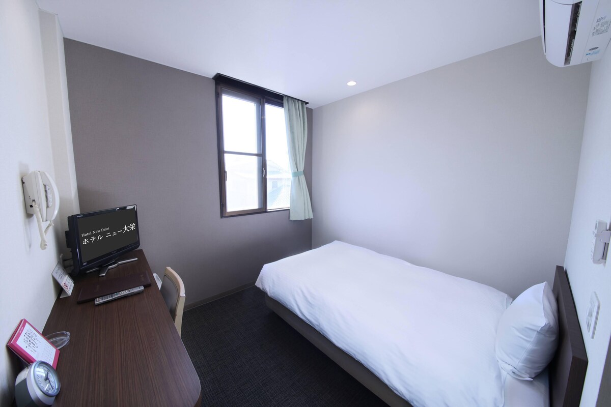 Habitación de hotel con una cama individual, un escritorio con computadora, teléfono y ventana con cortinas.