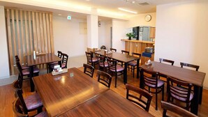 A dining area with wooden tables and chairs, a kitchen area with a refrigerator and stove, and a clock on the wall. - Business Hotel New Daiei (Ashikaga)