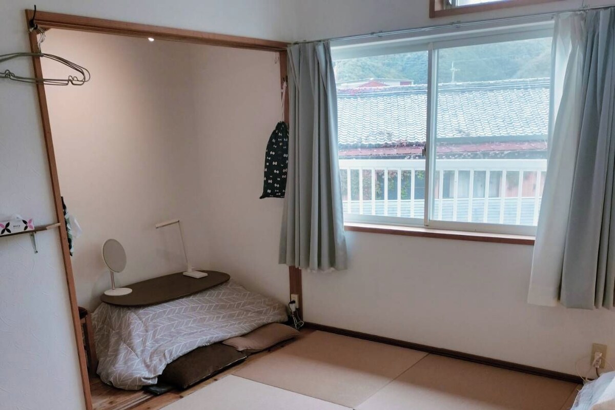A room with tatami flooring, a bed with a white and beige bedspread, a small bedside table with a mirror, and a window with curtains.