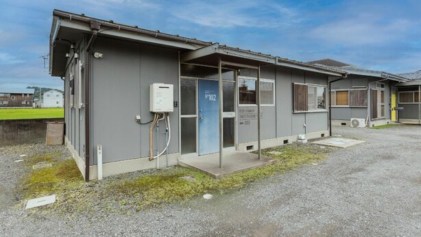 Un bâtiment à la façade grise, une porte bleue portant l’inscription « n° 102 », et un chauffe-eau fixé au mur.
