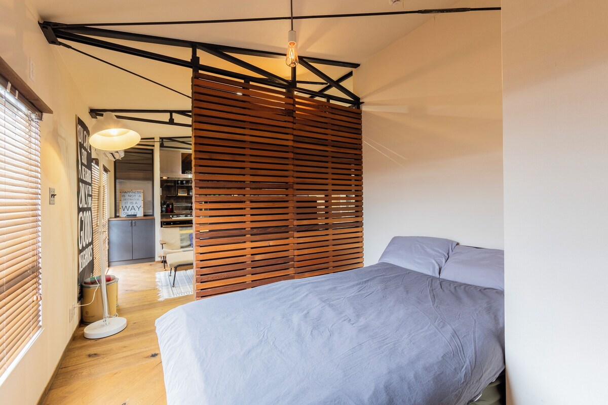 Ein Schlafzimmer mit einem Bett, Holzjalousien, einer Hängeleuchte und Blick in den Küchenbereich.