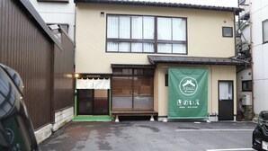 A building with a green awning displaying a logo and Japanese text, a small front porch, and a parked car. - Kinosaki Onsen Kinoie (Toyooka)