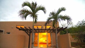 A modern building with a covered patio area, palm trees, and a visible bedroom with a bed and window. - Somos (Nakijin)