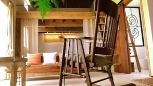 A rustic wooden rocking chair in a cozy room with a wooden bench, a sofa with cushions, and a large fern plant. - Somos (Nakijin)
