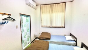 Een kleine, goed verlichte kamer met twee eenpersoonsbedden, een raam met gordijnen, een airconditioner, een ventilator en een deur met gordijn.