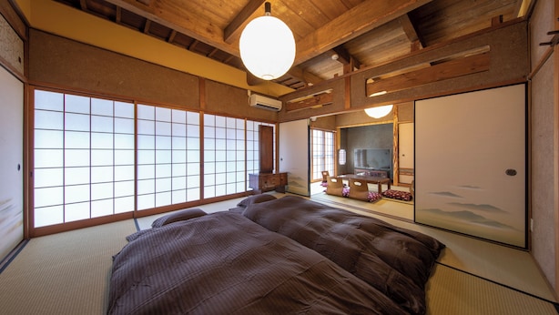 Une pièce de style japonais traditionnel, avec un sol recouvert de tatamis, un grand lit doté d’une couvre-lit marron foncé, des portes coulissantes et un plafond en bois.