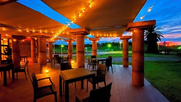 Un área de comedor al aire libre con mesas y sillas, cubierta por un toldo e iluminada con luces colgantes.