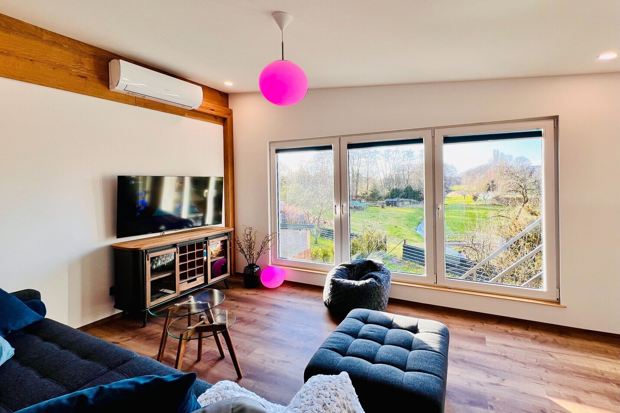 A modern living room with a large TV, wooden flooring, a black sofa, a glass table, and a view of a green landscape through large windows.