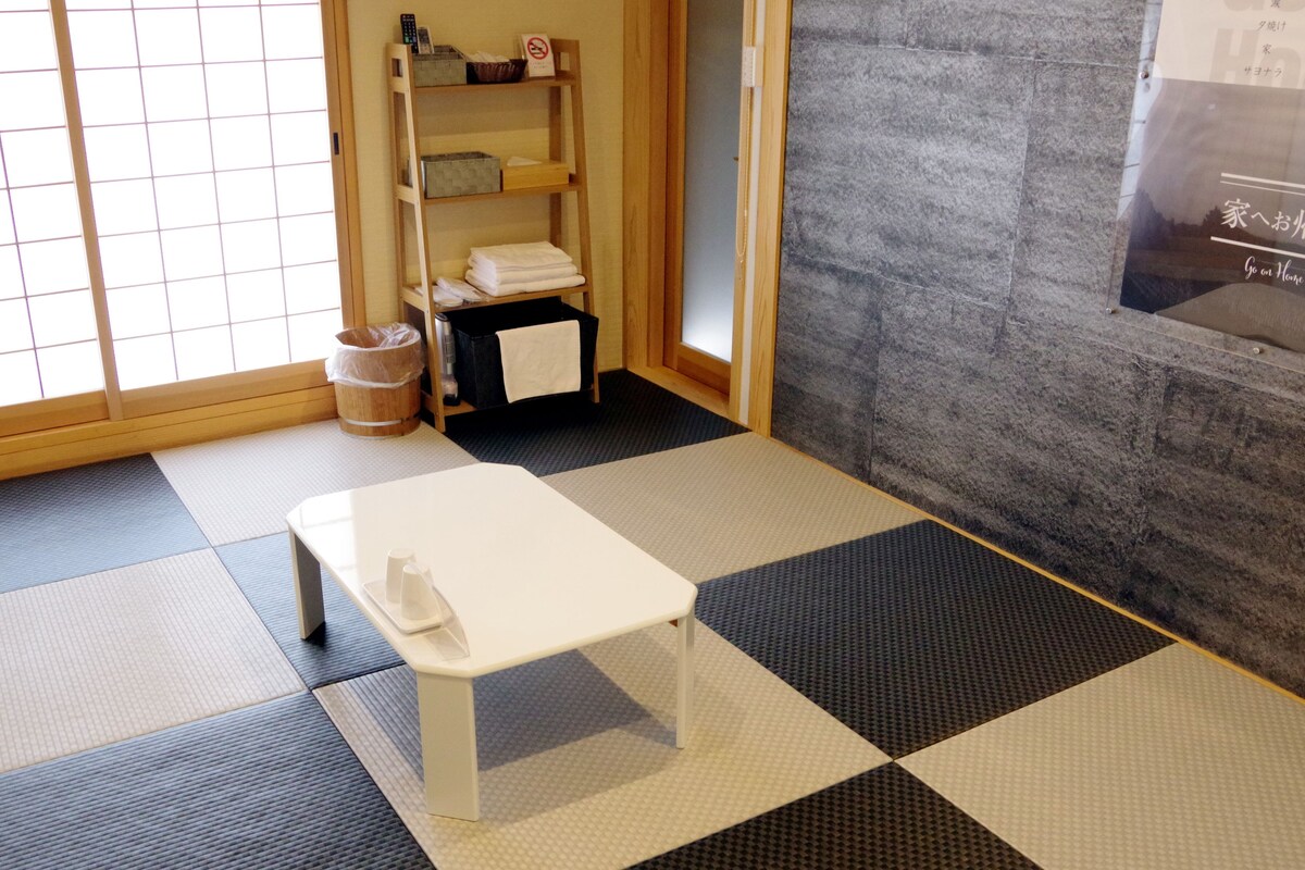 A room with tatami mat flooring, a small white table, a shelf with towels, and a stone wall.