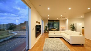 A modern living room with a large glass door leading to a balcony, a white sectional sofa, and a wooden floor. - Chichibugahama Resort Villa Senja (Mitoyo)